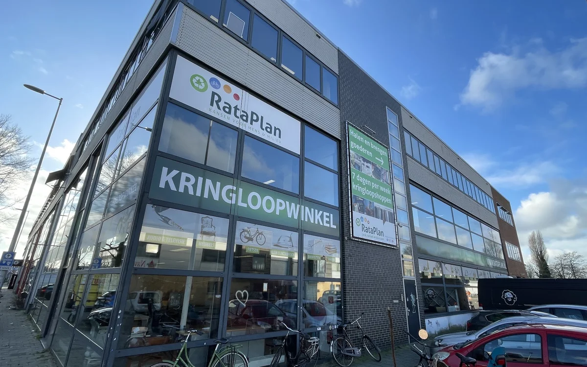 Gevel van kringloopwinkel RataPlan met grote ramen, winkelnaam duidelijk zichtbaar, enkele fietsen ervoor, rode auto rechts