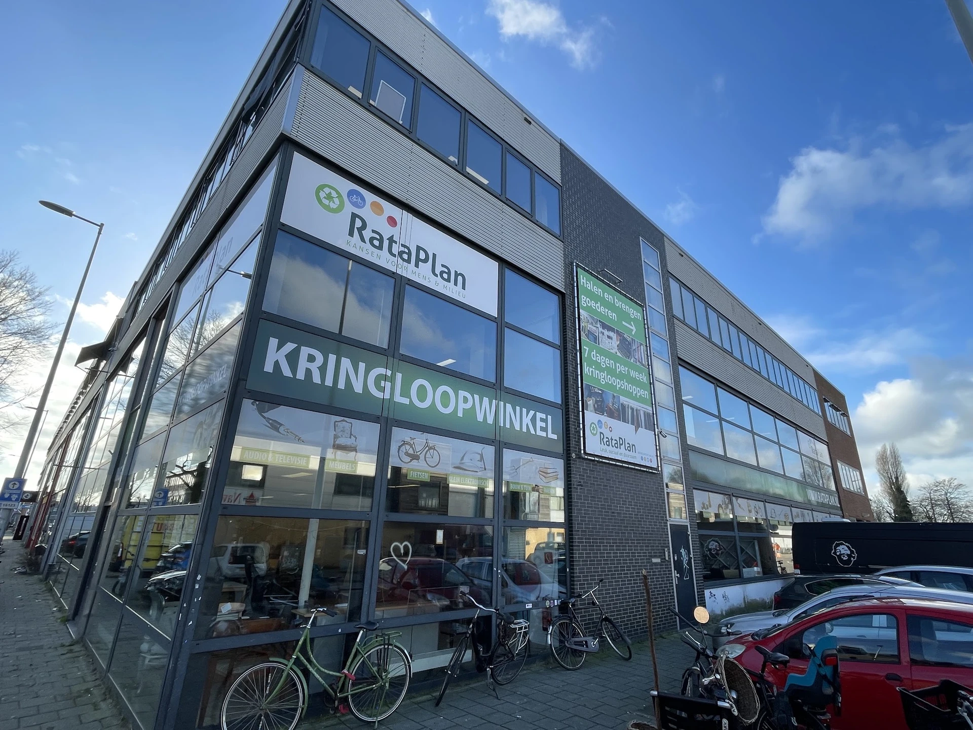 Gevel van kringloopwinkel RataPlan met grote ramen, winkelnaam duidelijk zichtbaar, enkele fietsen ervoor, rode auto rechts