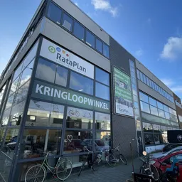 Gevel van kringloopwinkel RataPlan met grote ramen, winkelnaam duidelijk zichtbaar, enkele fietsen ervoor, rode auto rechts