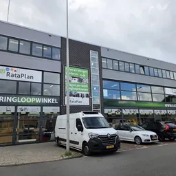 Gevel van kringloopwinkel met etalages en parkeerplaats ervoor, witte bestelbus geparkeerd