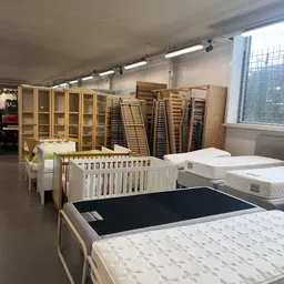 Uitgestalde meubels in een kringloopwinkel: witte babybedje, matrassen, kasten en houten lattenbodem met zwart frame