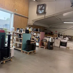 Binnenaanzicht van kringloopwinkel met diverse gebruikte huishoudapparatuur, meubels en kastjes uitgestald op de vloer en teg