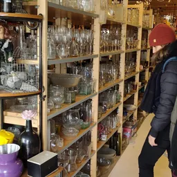 Uitgestalde glazen en drinkglazen in verschillende vormen en maten op houten stellingen in een kringloopwinkel