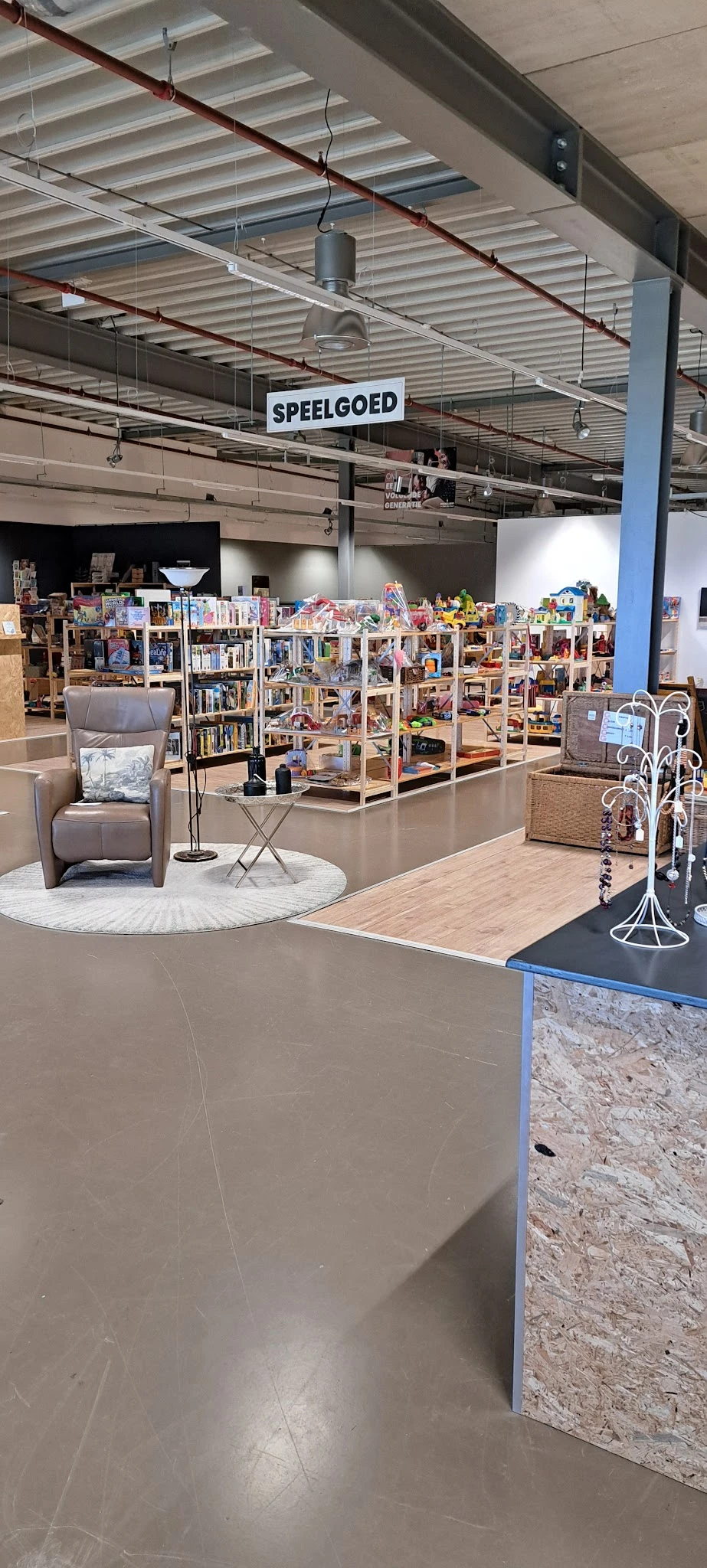 Winkelvloer met houten stellingen vol tweedehands spullen, grijze fauteuil op wit rond tapijt, sieradenstandaard en diverse d
