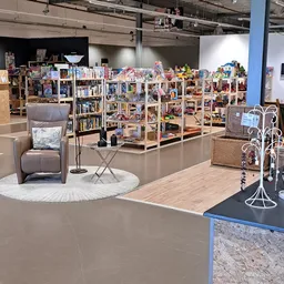 Winkelvloer met houten stellingen vol tweedehands spullen, grijze fauteuil op wit rond tapijt, sieradenstandaard en diverse d