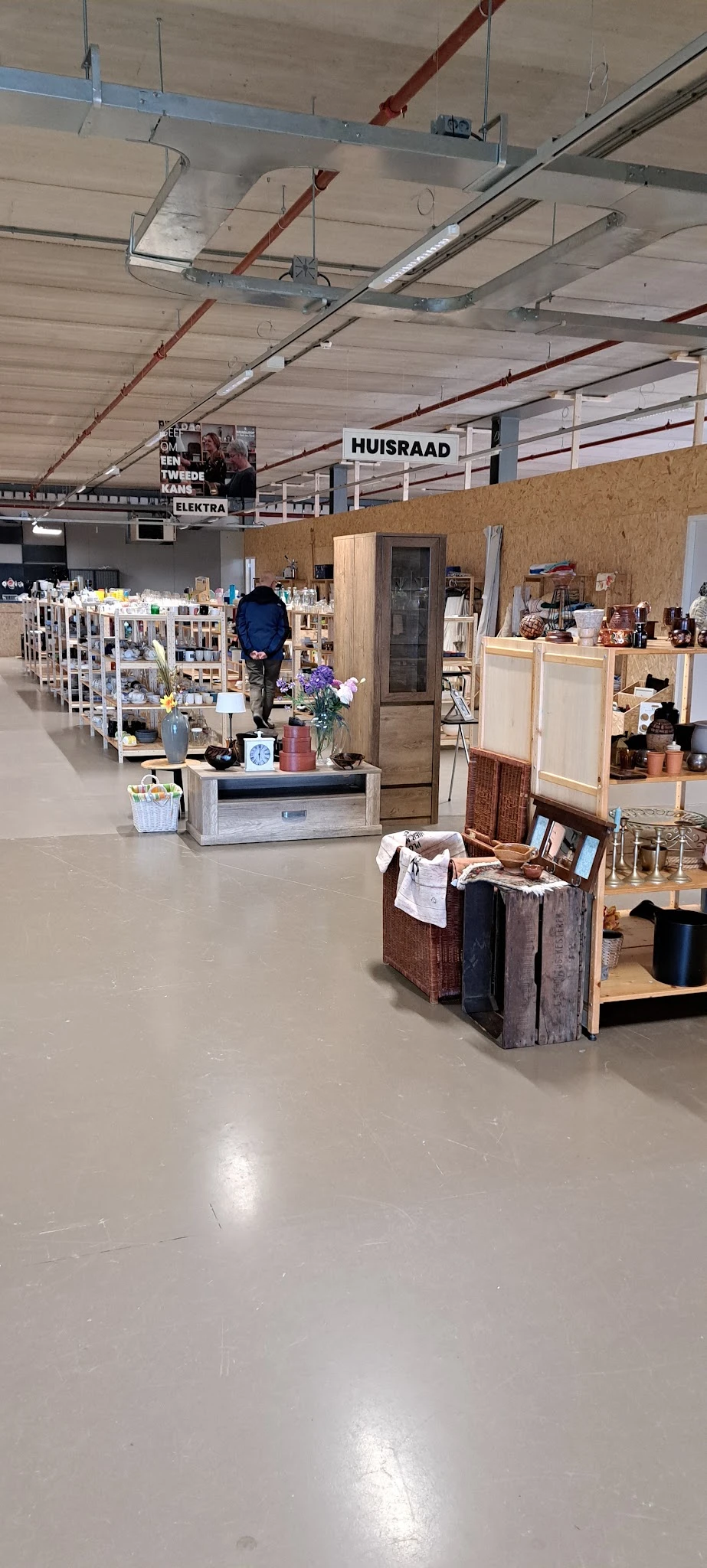 Winkelvloer van kringloopwinkel met diverse stellingen, huishoudartikelen, decoraties en een bezoeker die rondkijkt
