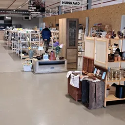 Winkelvloer van kringloopwinkel met diverse stellingen, huishoudartikelen, decoraties en een bezoeker die rondkijkt
