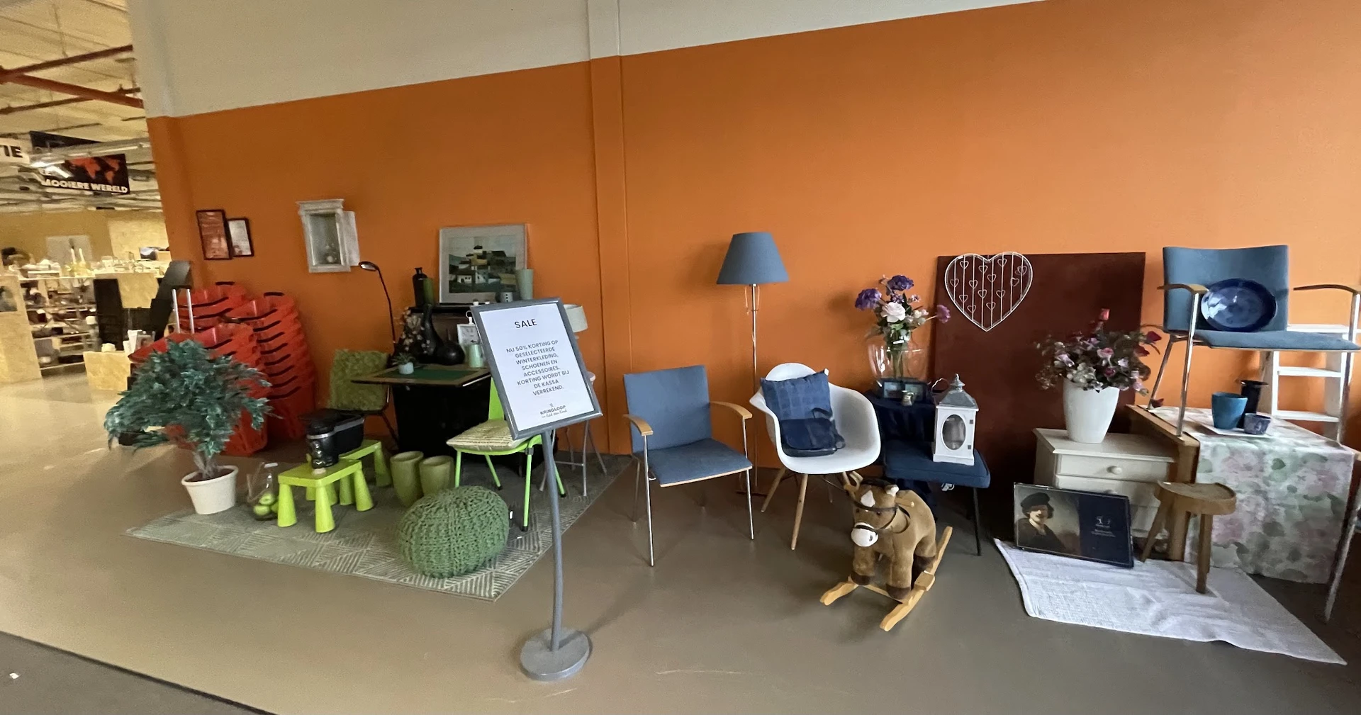 Winkelvloer met mogelijk kindermeubilair en speelgoed, oranje wand met verschillende stoelen, lampen en huishoudelijke artike