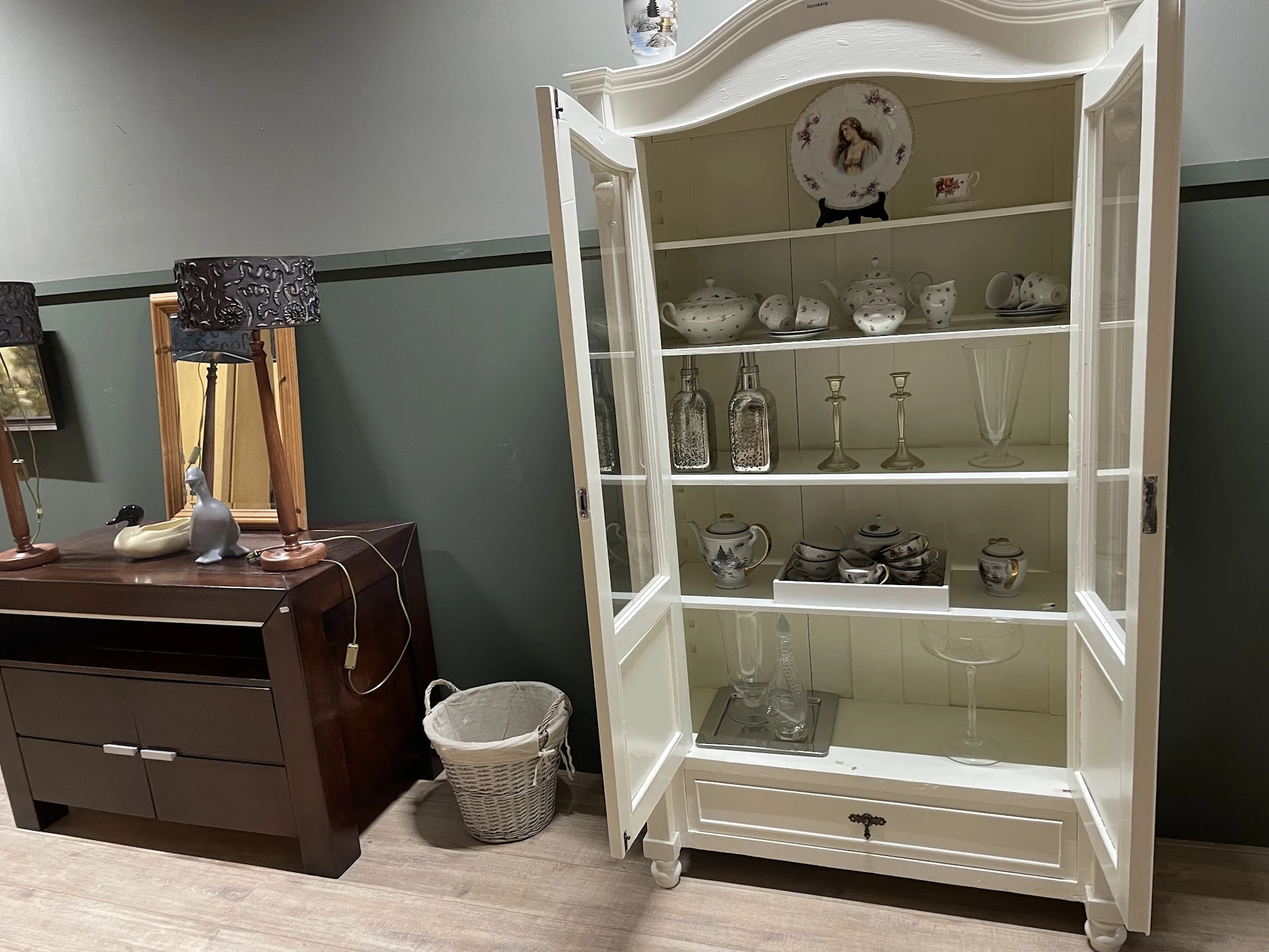 Witte glazen vitrinekast met decoratieve items zoals servies, glazen en flesjes, naast een donkerbruin meubelstuk