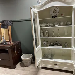 Witte glazen vitrinekast met decoratieve items zoals servies, glazen en flesjes, naast een donkerbruin meubelstuk