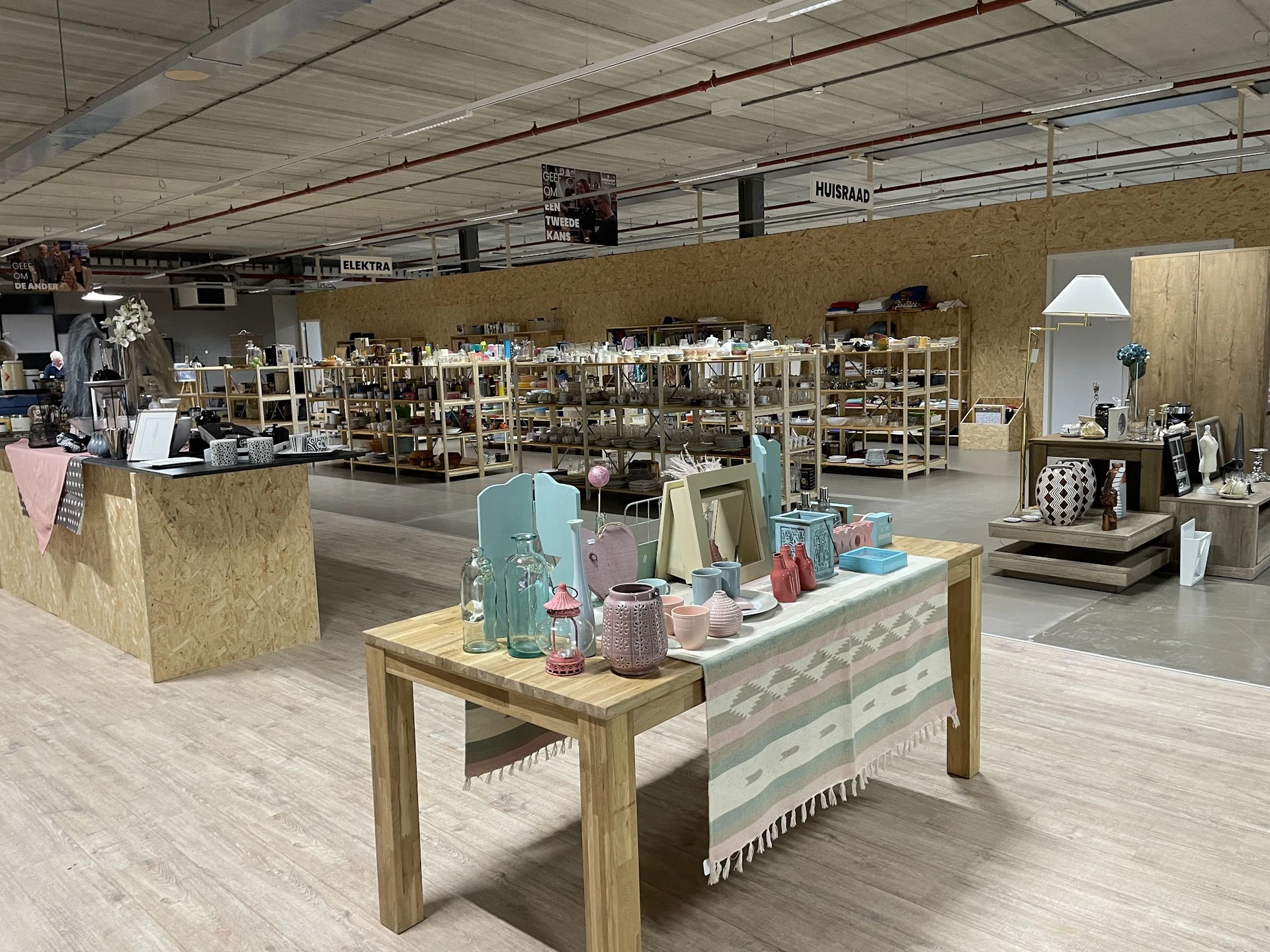 Ruime winkelvloer met houten toonbank en display met huishoudelijke artikelen, op achtergrond stellingen met meubels en inric