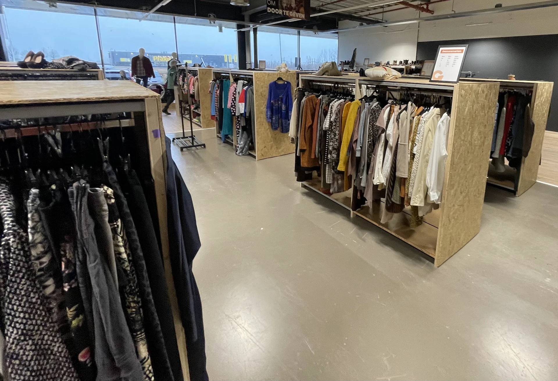 Winkelvloer met houten stellingen vol kleding aan hangers, grote ramen met uitzicht