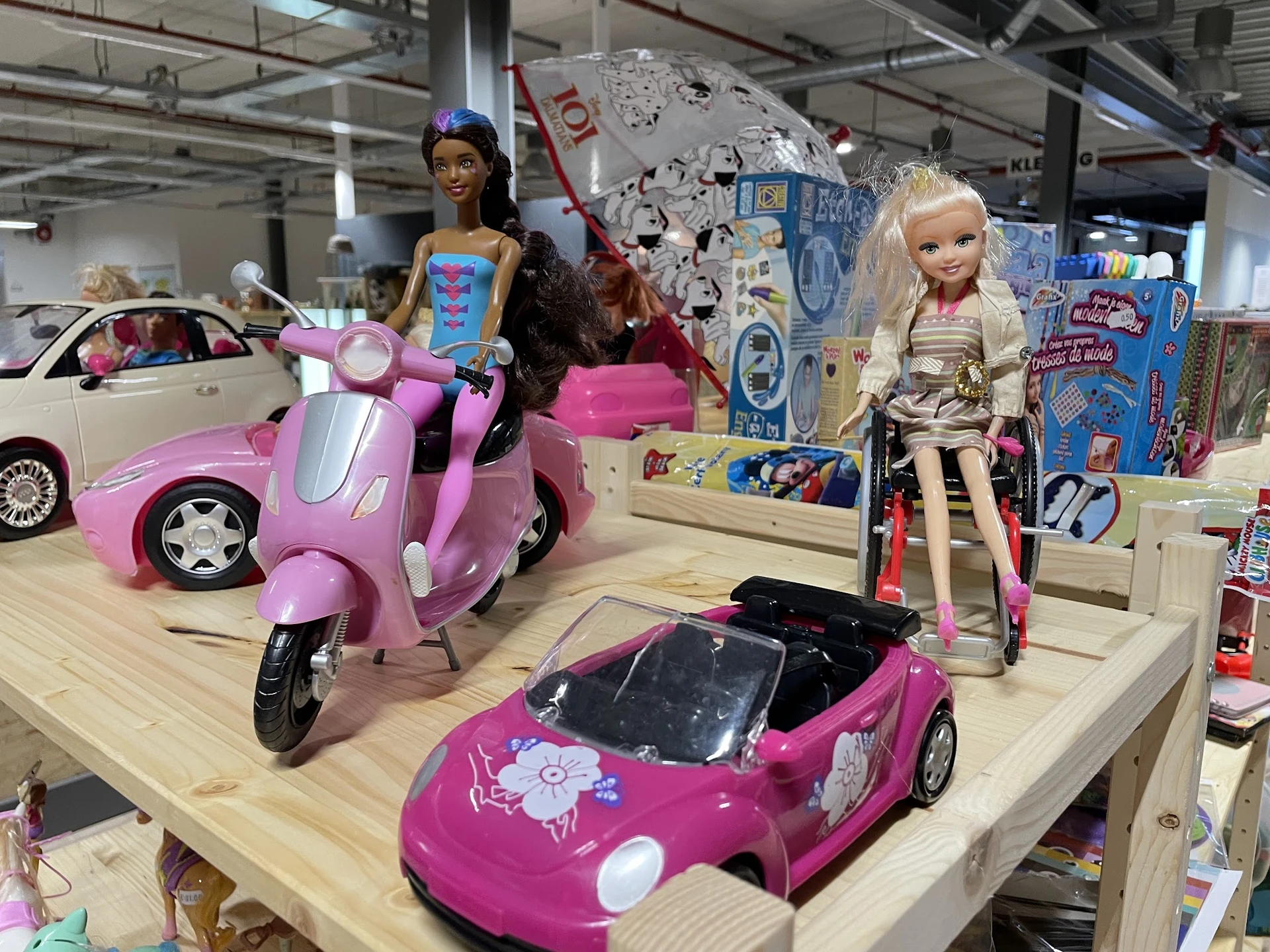 Uitgestalde speelgoed in de winkel, waaronder roze Barbie-voertuigen, een scooter en poppen met accessoires