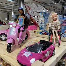 Uitgestalde speelgoed in de winkel, waaronder roze Barbie-voertuigen, een scooter en poppen met accessoires