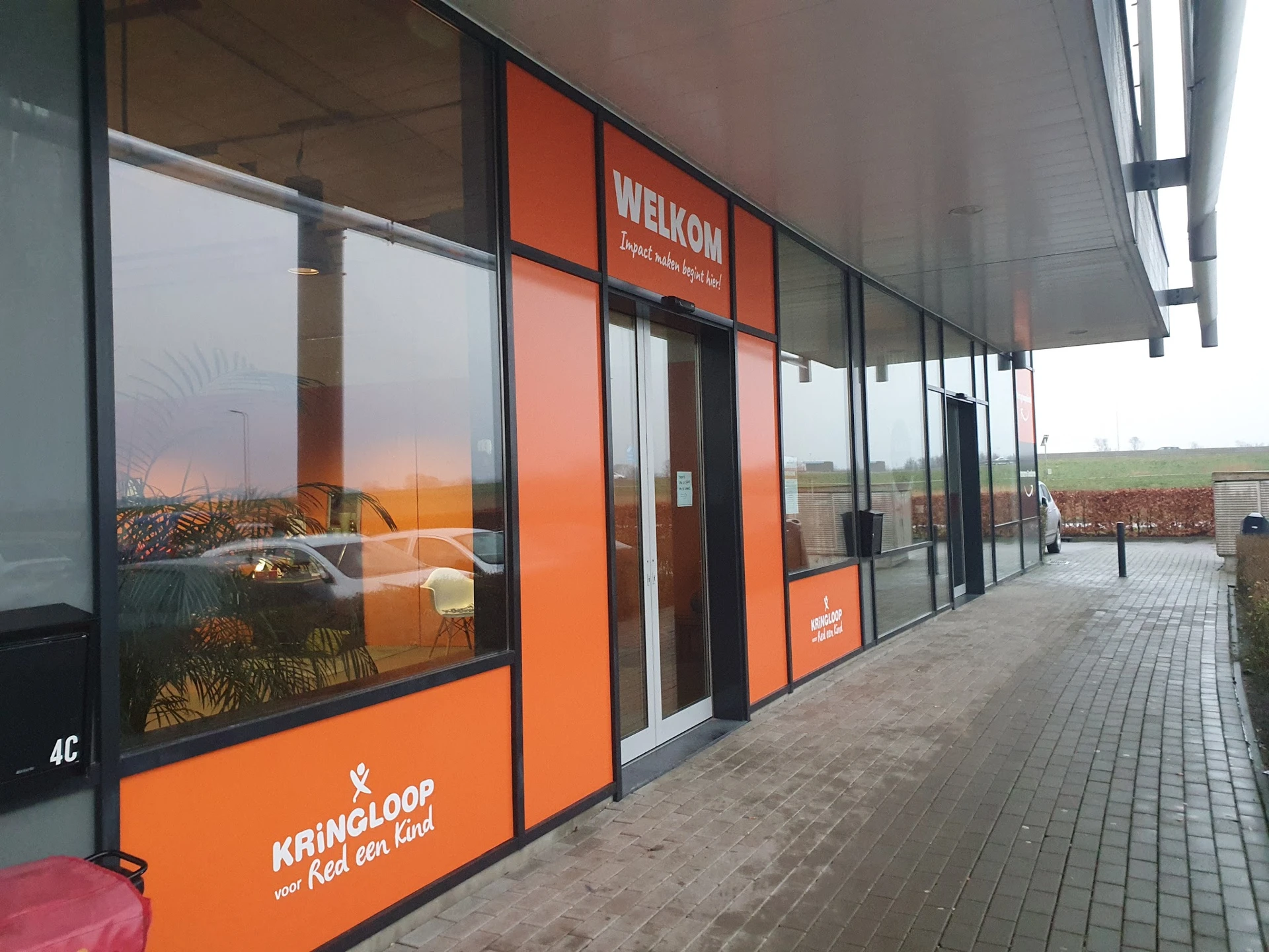 Winkelvoorgevel met oranje en zwarte ramen en deur, winkelnaam WELKOM zichtbaar, in een winkelstrip met plaveisel ervoor