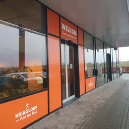 Winkelvoorgevel met oranje en zwarte ramen en deur, winkelnaam WELKOM zichtbaar, in een winkelstrip met plaveisel ervoor