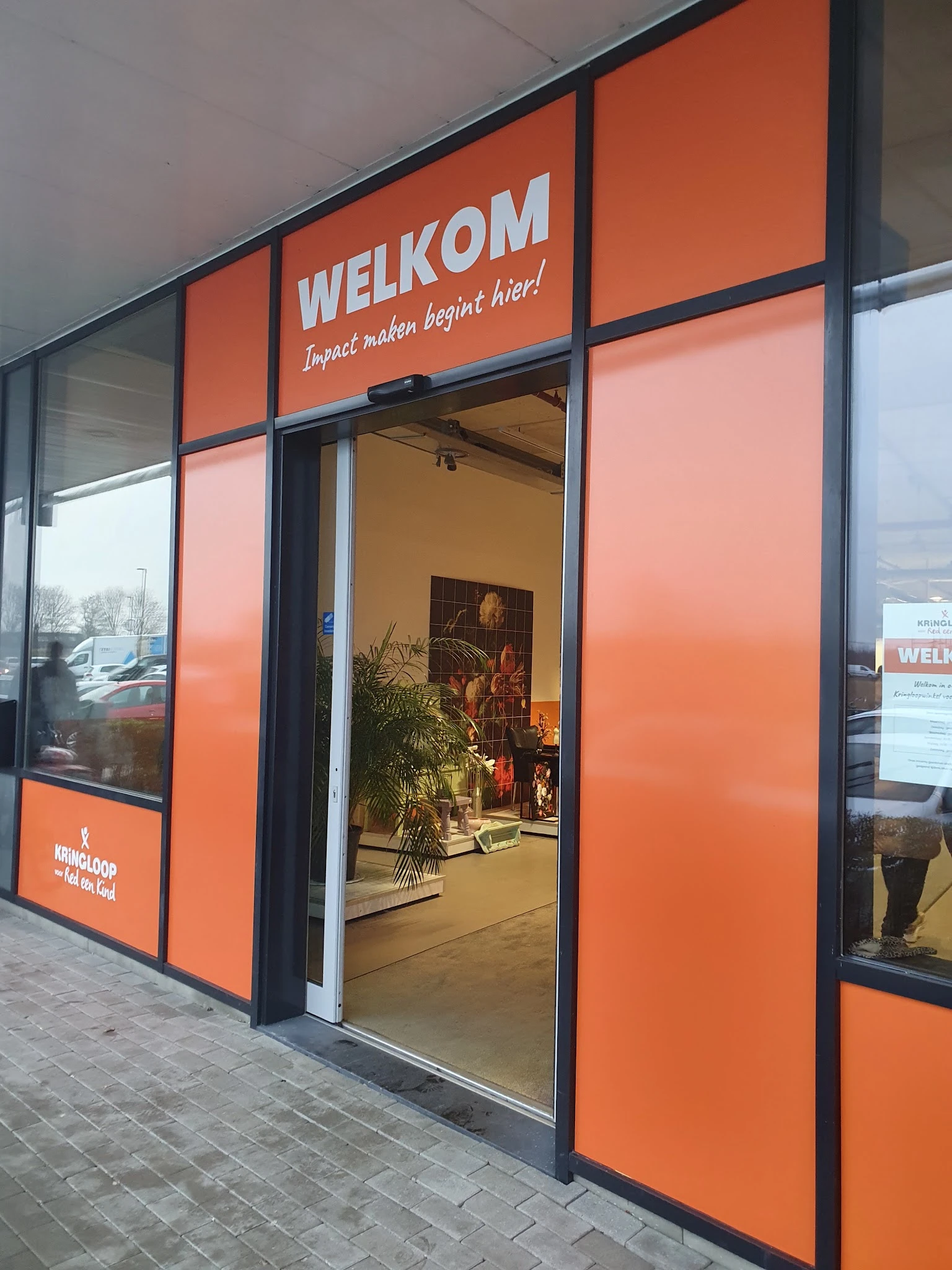 Winkelvoorgevel met grote oranje ramen en glazen deur, winkelnaam en slogan zichtbaar boven, plantjes en decoratie in etalage