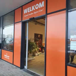 Winkelvoorgevel met grote oranje ramen en glazen deur, winkelnaam en slogan zichtbaar boven, plantjes en decoratie in etalage