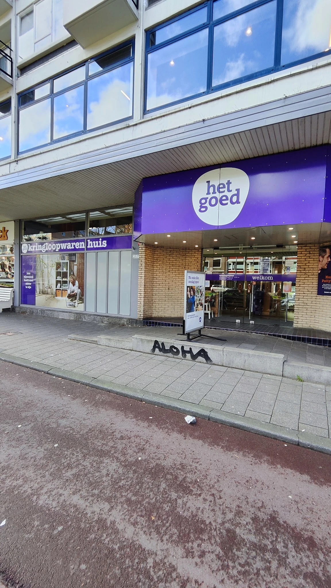 Gevel van kringloopwinkel met paarse gevel, wit logo 'het goed', winkelruit en ingang aan straat