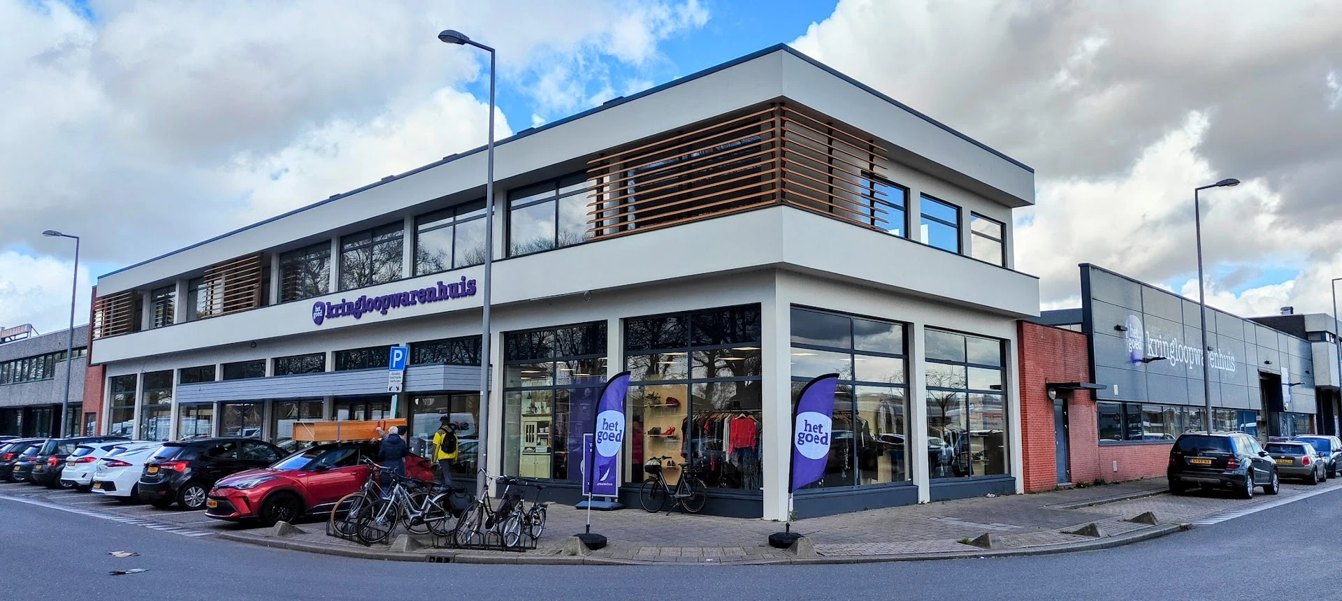 Moderne witte gevel van kringloopwinkel met grote ramen, houten luifels en blauwe accenten, fietsenstalling ervoor