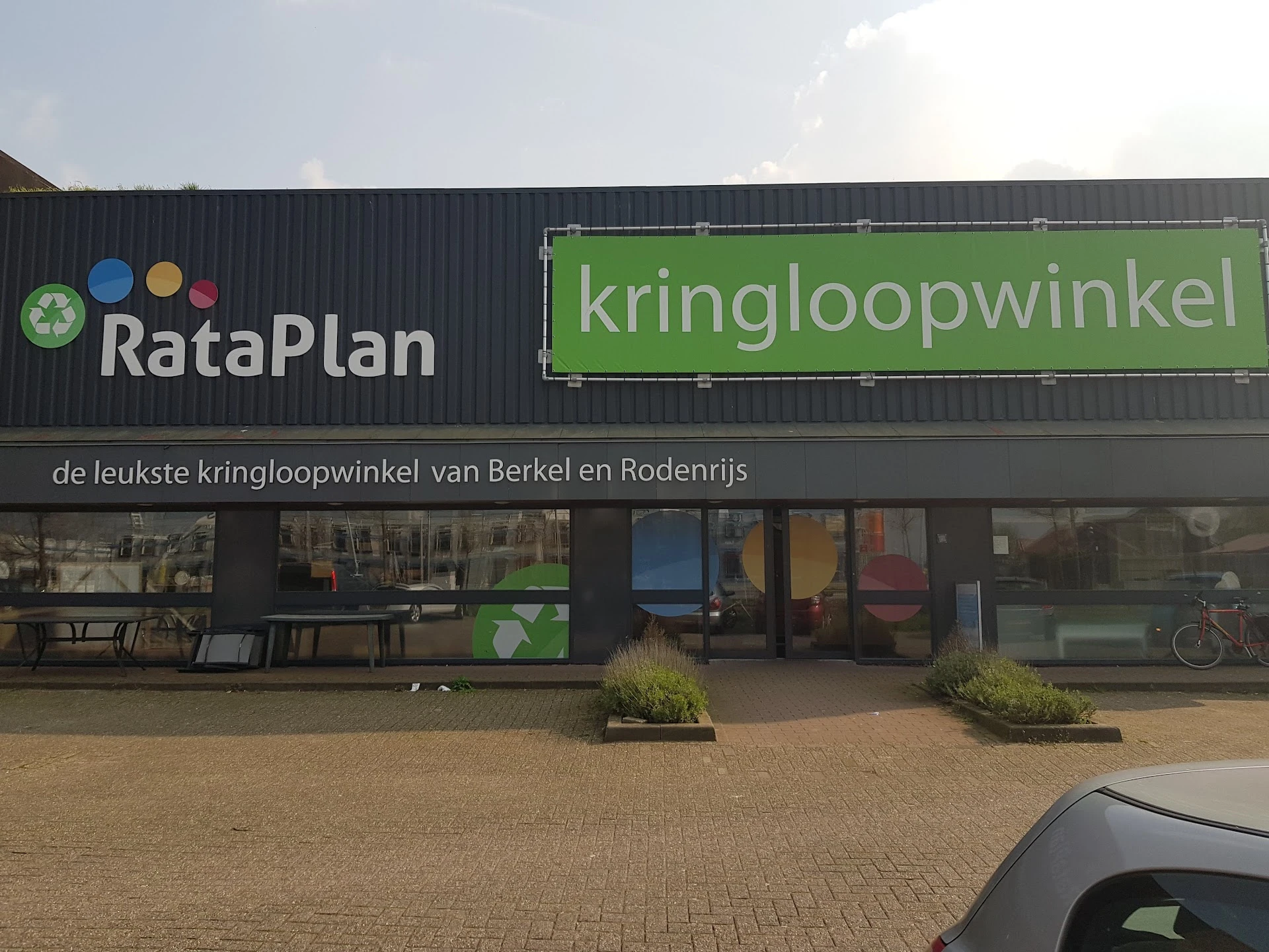 Gevel van kringloopwinkel RataPlan met groene kringloopwinkel-bord, donkergrijs pand met ramen en plantenbakken ervoor