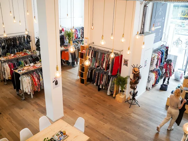 Ruim interieur van kringloopwinkel met kledingrekken, houten vloer, witte zuilen en hangende gouden lampen