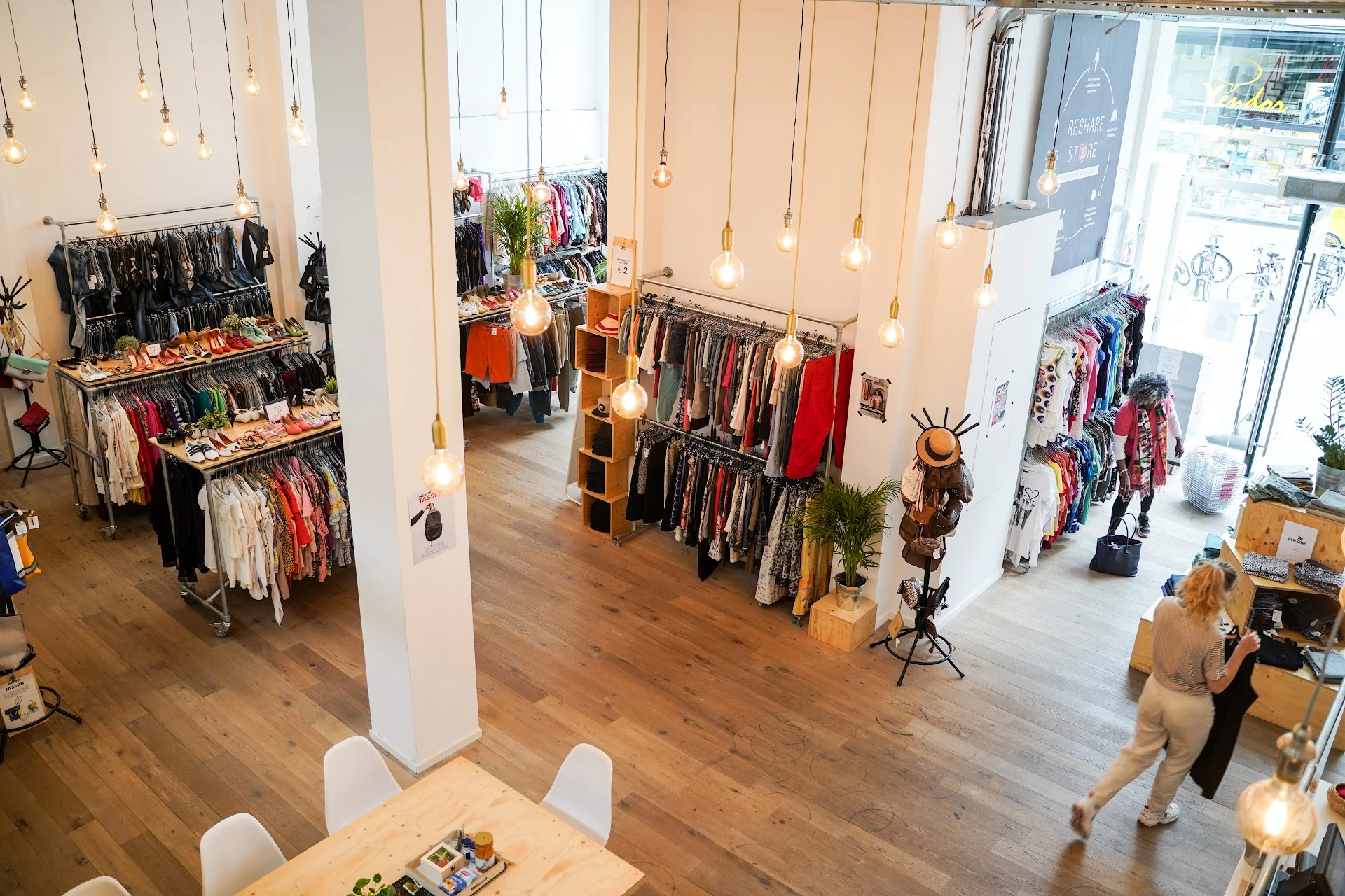 Ruim interieur van kringloopwinkel met kledingrekken, houten vloer, witte zuilen en hangende gouden lampen