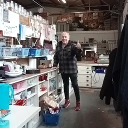 Man in geruite jas glimlachend in werkruimte van kringloopwinkel, omgeven door stellingen met spullen