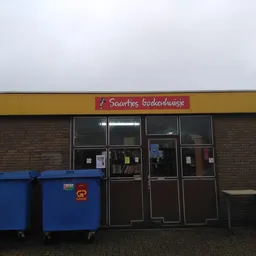 Gevel van kringloopwinkel met geel/bruine voorgevel, zwarte deuren en blauwe afvalcontainers ervoor