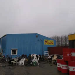 Blauwe magazijnbouw met witte ramen en gele afdak, omgeven door gebruikt meubilair en rode vaten buiten