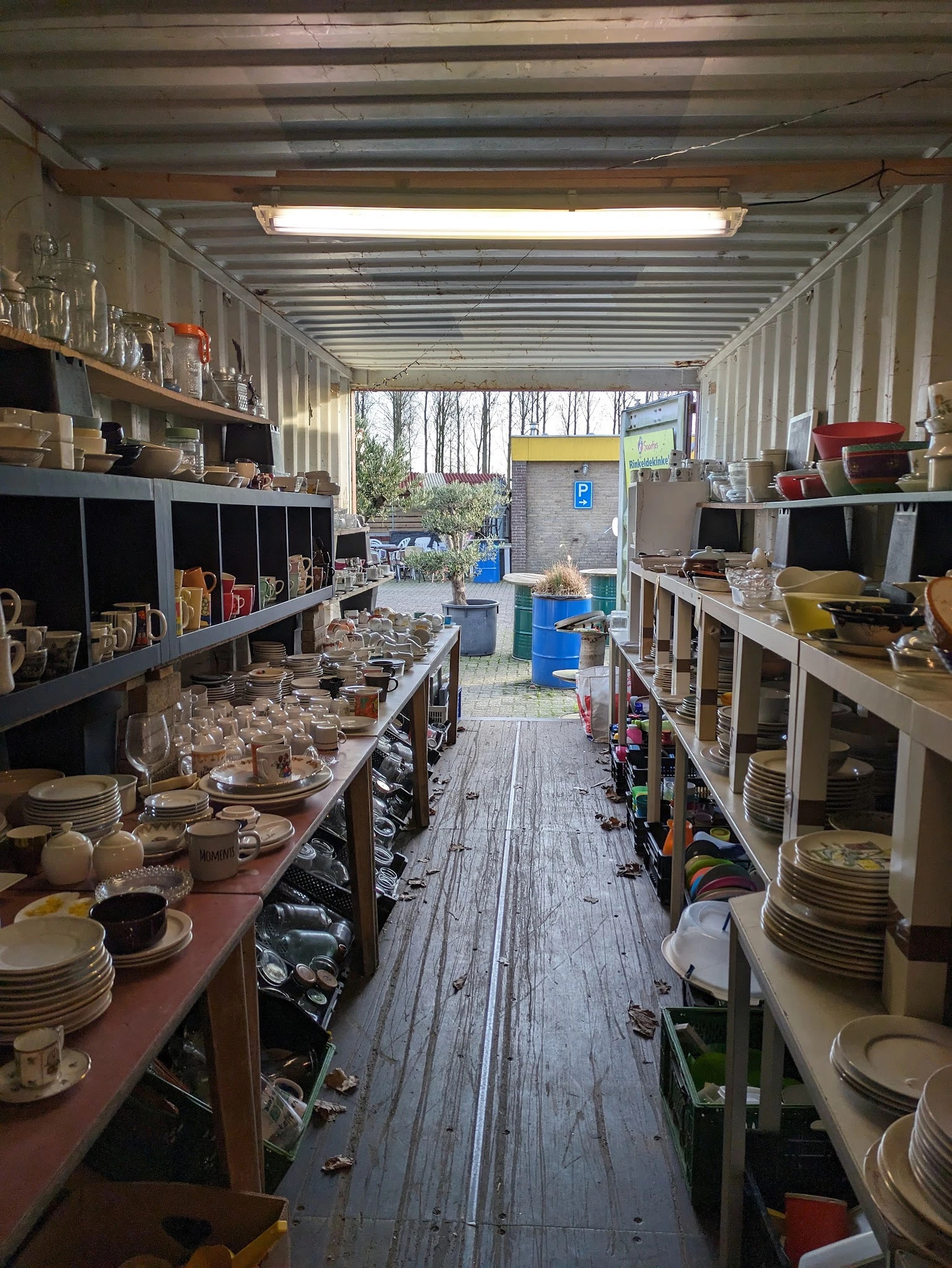 Binnenaanzicht van kringloopwinkel met stellingen vol serviesgoed, glazen en huishoudelijke artikelen langs beide zijden