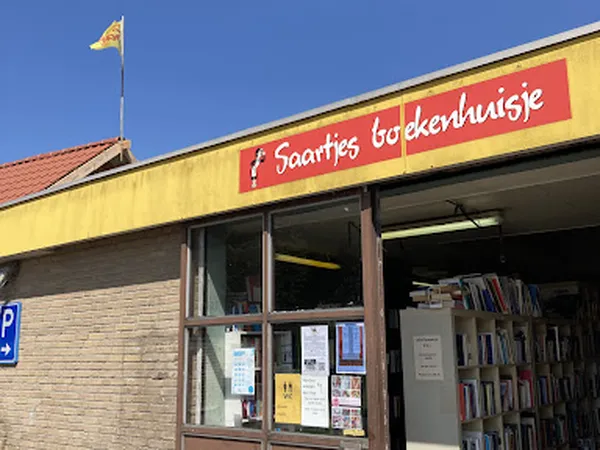 Gele gevel van kringloopwinkel met rode naam 'Saartje boekenhuisje', parkeerplaats en Nederlandse vlag op dak
