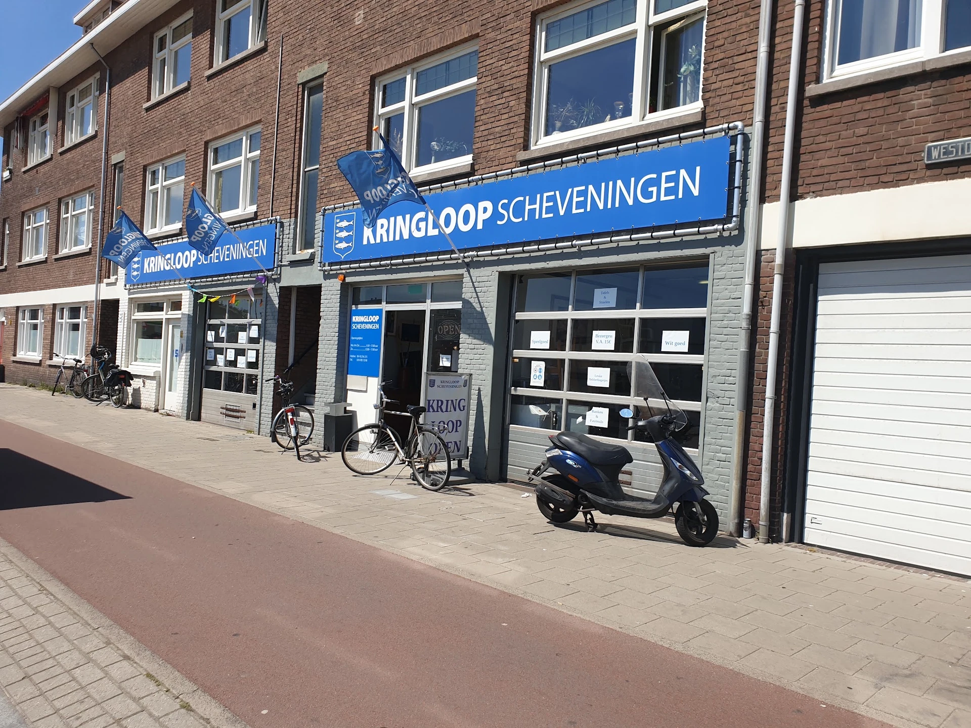 Gevel van kringloopwinkel met blauwe uithangbord 'Kringloop Scheveningen', fiets en scooter ervoor