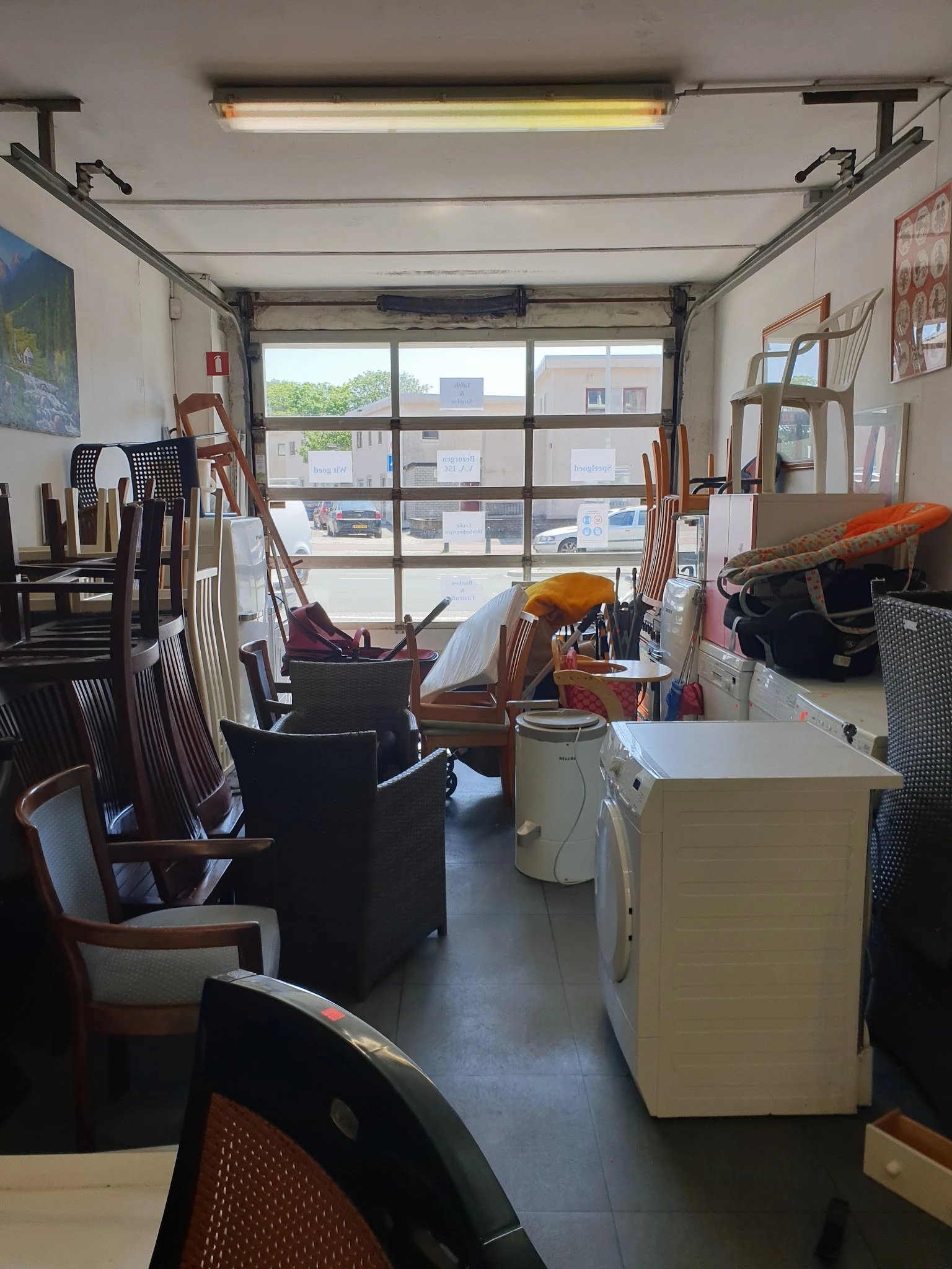 Binnenaanzicht van kringloopwinkel met stoelen, tafels en diverse spullen langs ramen met grote glazen pui