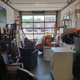 Binnenaanzicht van kringloopwinkel met stoelen, tafels en diverse spullen langs ramen met grote glazen pui