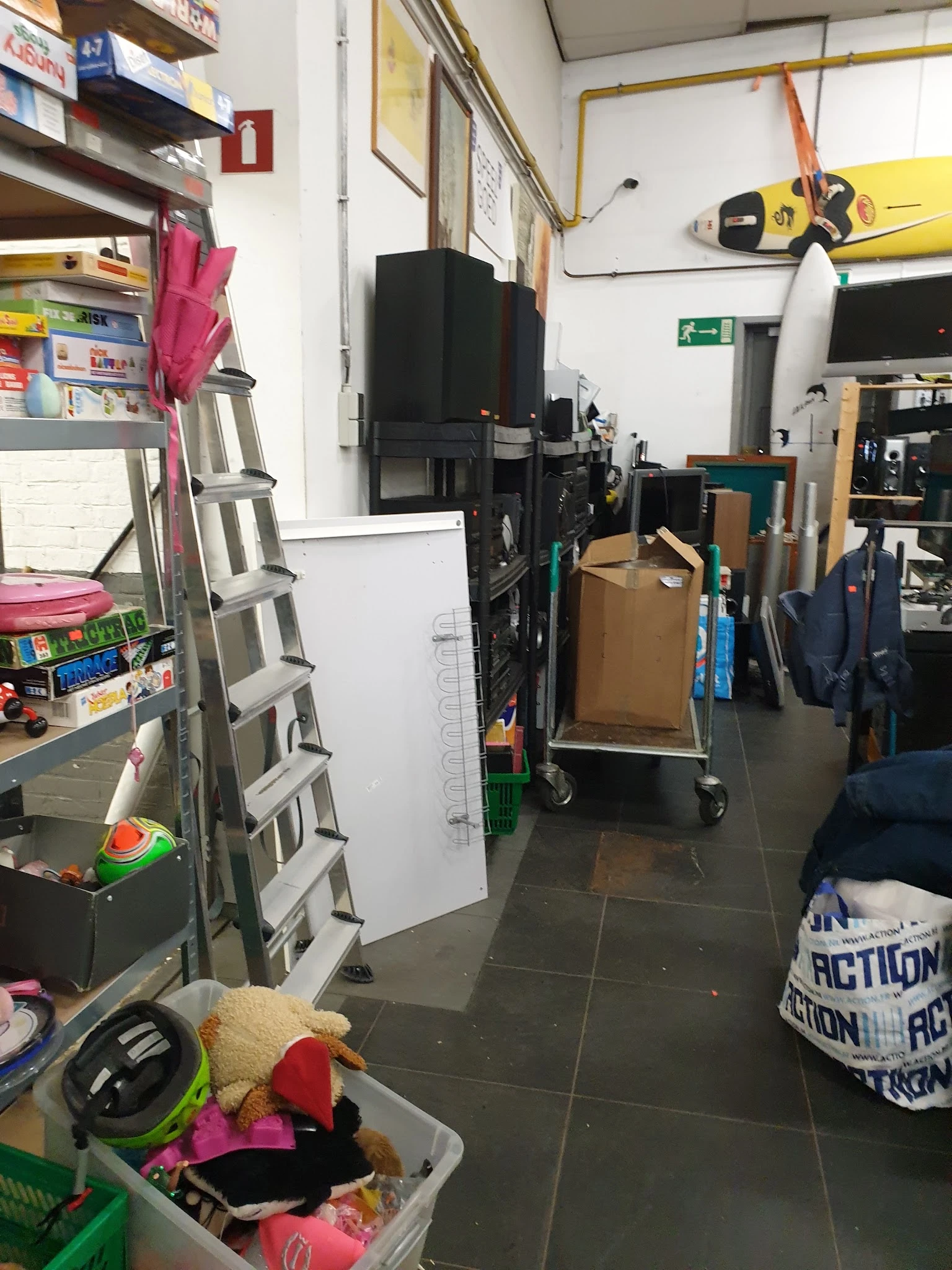 Winkelgang met stellingen, een ladder, kartonnen dozen op een kar en diverse spullen langs de muur