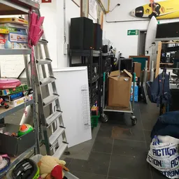 Winkelgang met stellingen, een ladder, kartonnen dozen op een kar en diverse spullen langs de muur