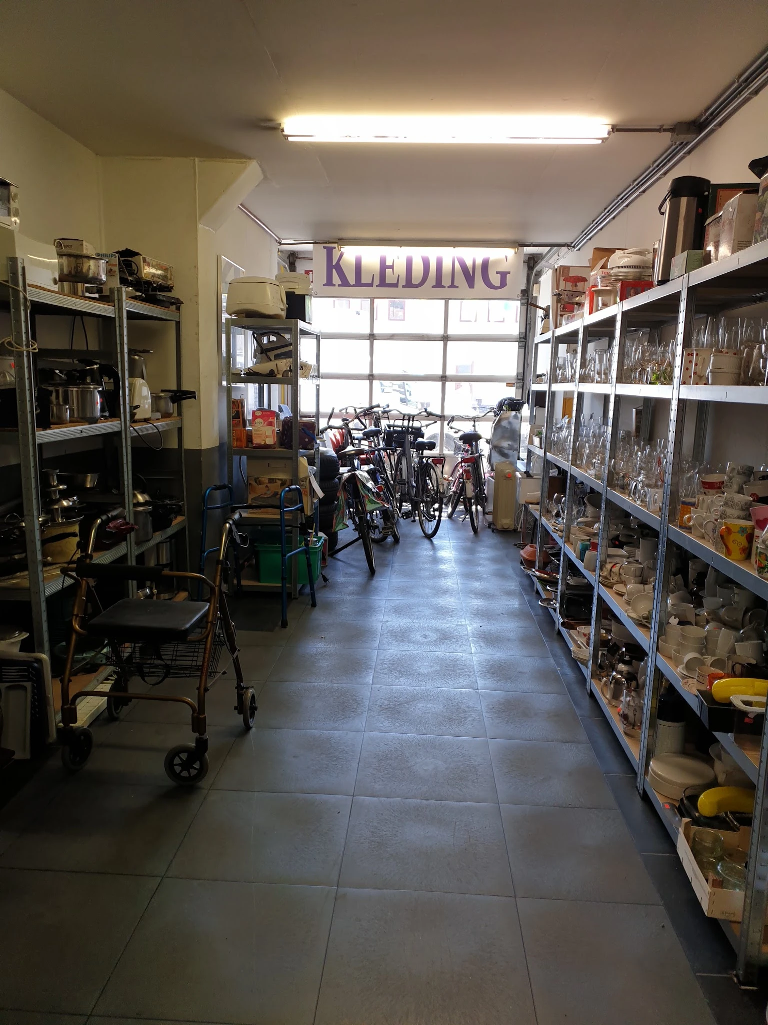 Winkelvloer van kringloopwinkel met stellingen vol spullen aan beide zijden, fietsen in het midden en 'KLEDING' bord boven