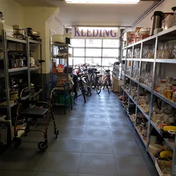 Winkelvloer van kringloopwinkel met stellingen vol spullen aan beide zijden, fietsen in het midden en 'KLEDING' bord boven