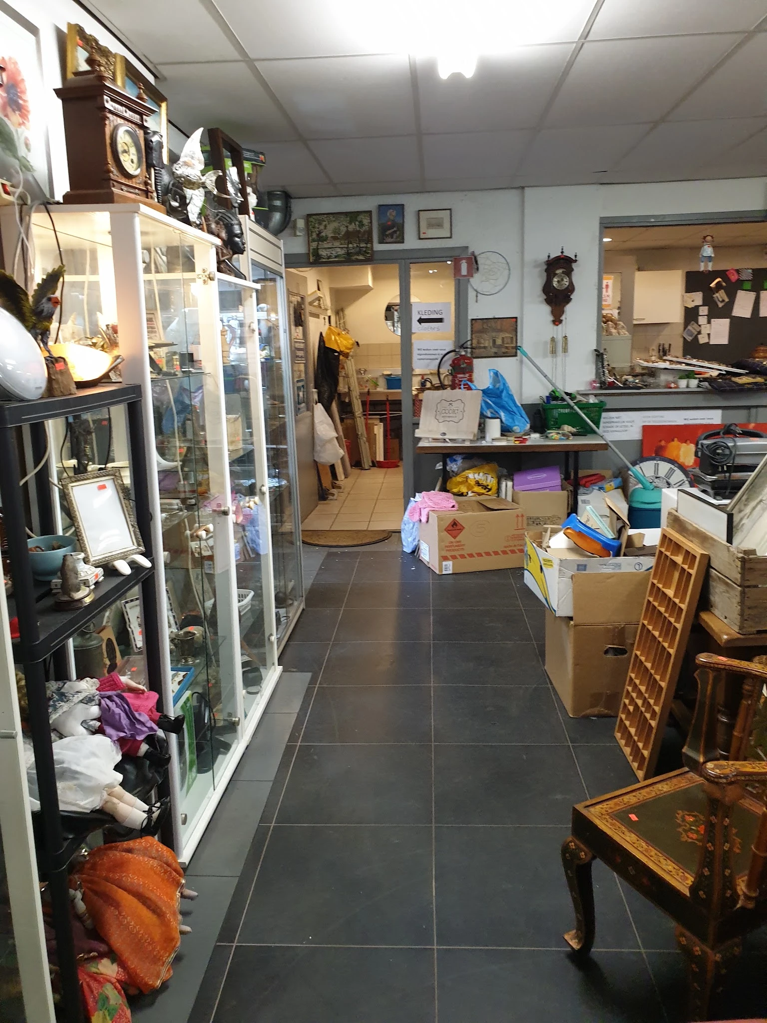 Binnenaanzicht van kringloopwinkel met stellingen, dozen, kleding en huishoudartikelen langs de muren en op de vloer.