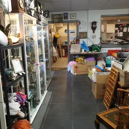 Binnenaanzicht van kringloopwinkel met stellingen, dozen, kleding en huishoudartikelen langs de muren en op de vloer.