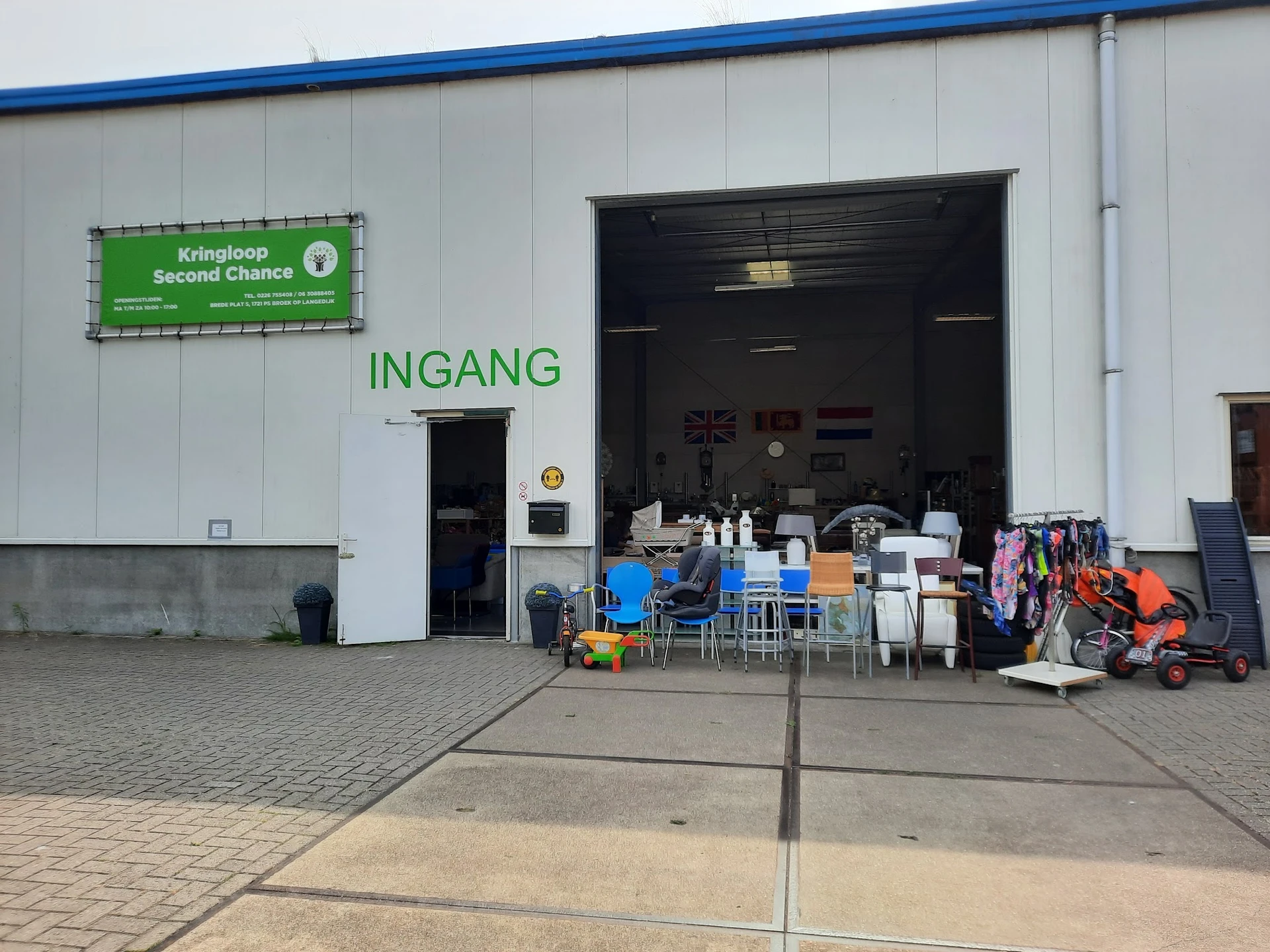 Voorkant van kringloopwinkel met groene naambordje en open garage-achtige deur, diverse gebruikte goederen buiten uitgestald
