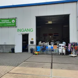 Voorkant van kringloopwinkel met groene naambordje en open garage-achtige deur, diverse gebruikte goederen buiten uitgestald
