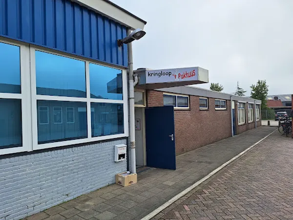 Voorgevel van kringloopwinkel met blauwe golfplaten dak, grote ramen, bakstenen muur en blauwe deur, winkaluifel met naam zic