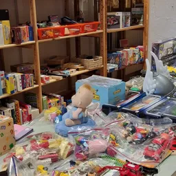 Speelgoed en boeken uitgestald op tafels voor de winkelschappen, waaronder knuffelbeesten, spellen en kinderboeken