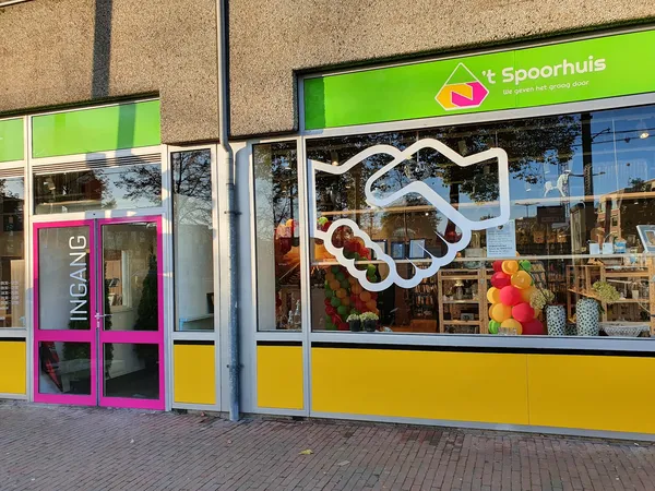 Voorgevel van kringloopwinkel 't Spoorhuis met groene bovenbalk, gele onderpui, roze deurkozijn en decoratieve witte handslip