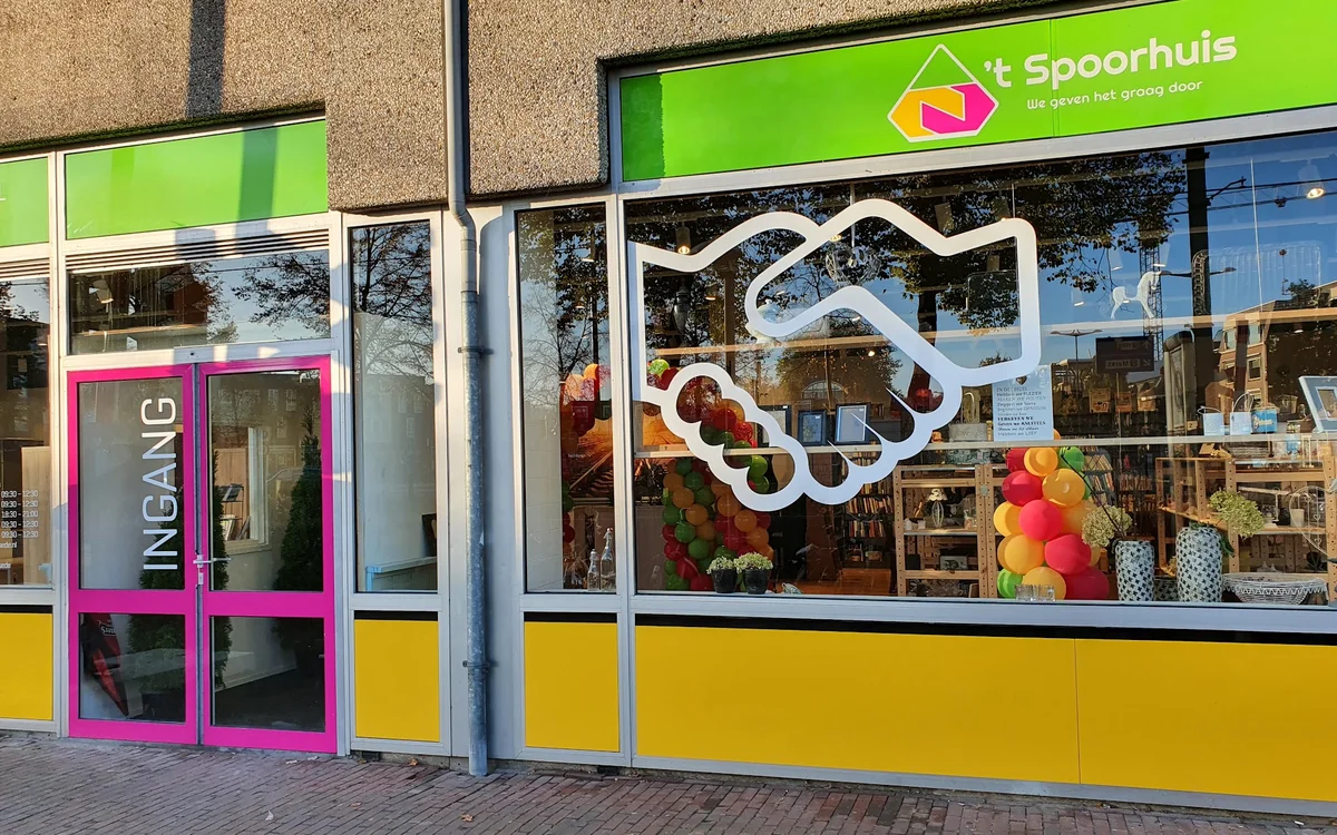 Gevel van kringloopwinkel 't Spoorhuis met groen en geel-blauw geschilderde pui, grote ramen en decoratieve witte slinger in 