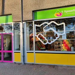 Voorgevel van kringloopwinkel 't Spoorhuis met groene bovenbalk, gele onderpui, roze deurkozijn en decoratieve witte handslip