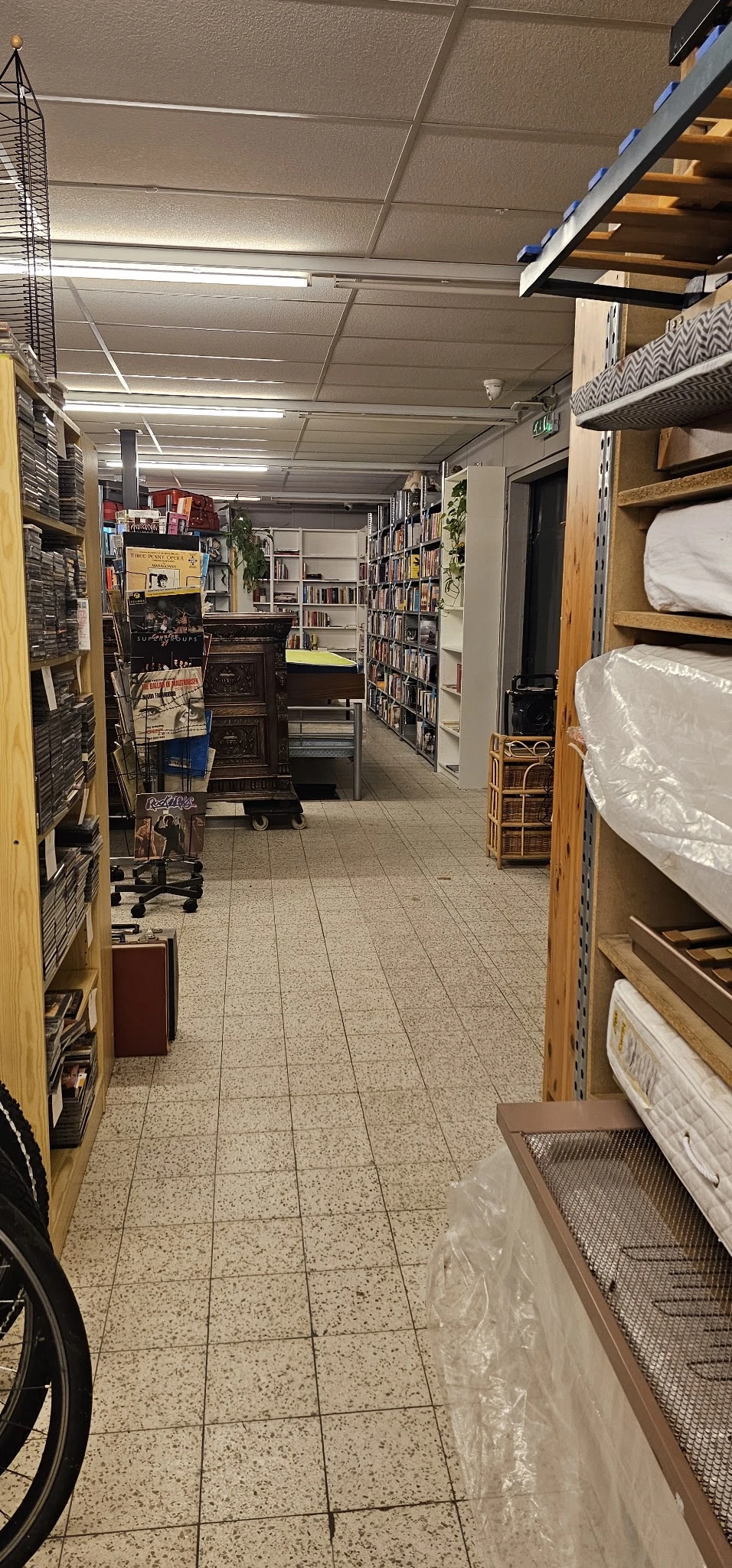 Winkelinterieur met boekenrekken, een donkere houten kast, bedden met matrassen en diverse huisraad langs de wanden