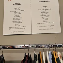Prijslijst met kledingstukken en schoenen, onder een rek met hangers met kleding